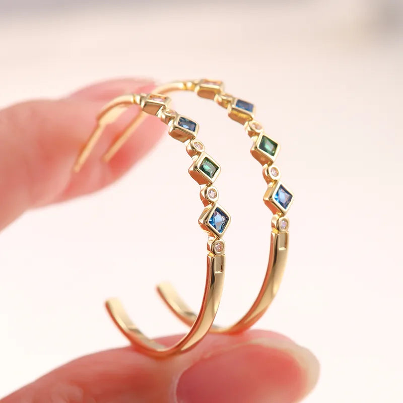 Gold hoop earrings with multicolor square gemstones, held between fingers