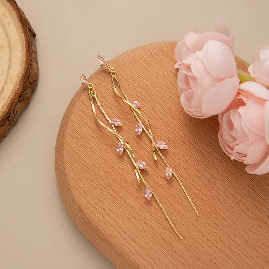 Gold-tone leaf drop earrings with pink crystal accents on wooden surface next to pink flowers