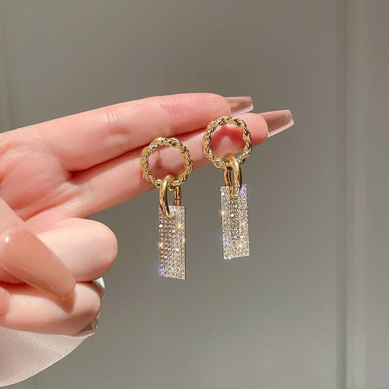Gold hoop earrings with textured rectangular crystal dangles, held by a hand