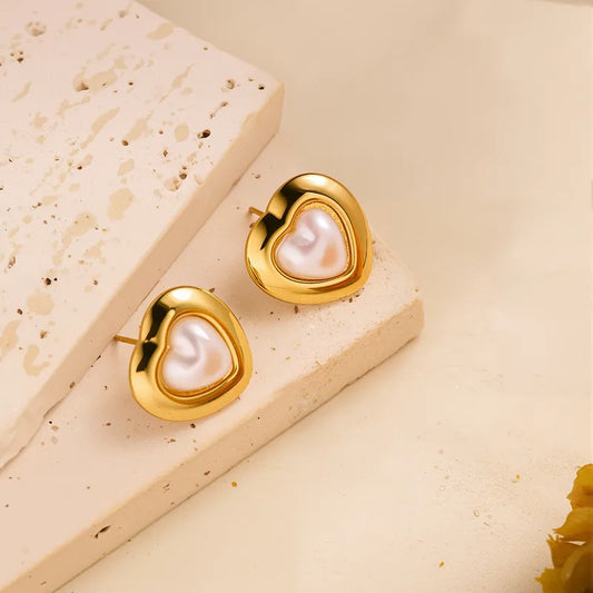 Gold heart-shaped stud earrings with pearl centers on a beige stone background